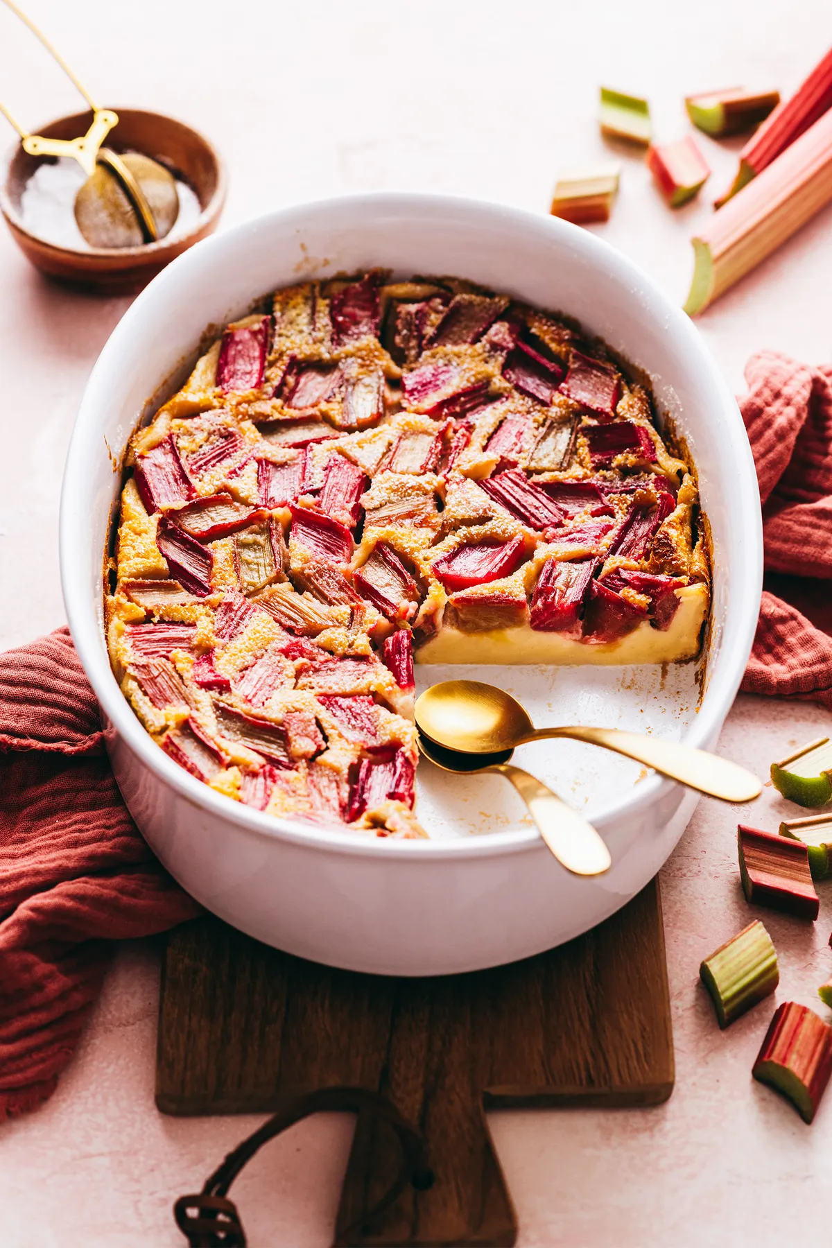 Tranche de clafouti à la rhubarbe sur assiette blanche, bords dorés et éclats de compote rosée, tige de rhubarbe en arrière-plan