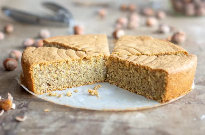 Un gâteau rond doré, tranché pour révéler une mie noisette claire, nappé d'un léger voile de praliné brillant et quelques noisettes torréfiées entières en décoration