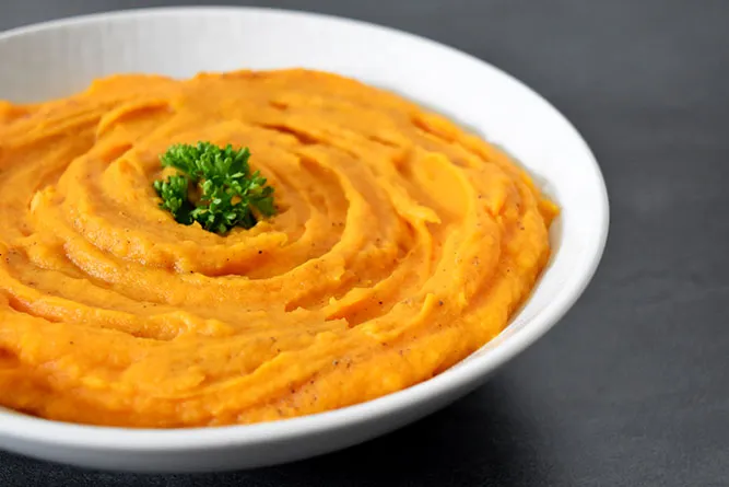 Bol en céramique contenant une purée de patate douce orange vif, nappée d'un filet d'huile d'olive et parsemée de graines et d'herbes fraîches